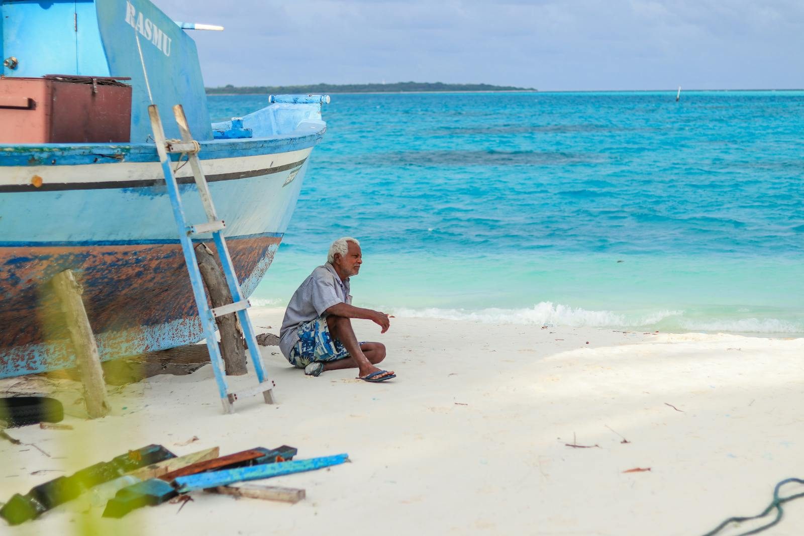 Elderly man resting by a fishing boat on a serene tropical beach with turquoise waters.